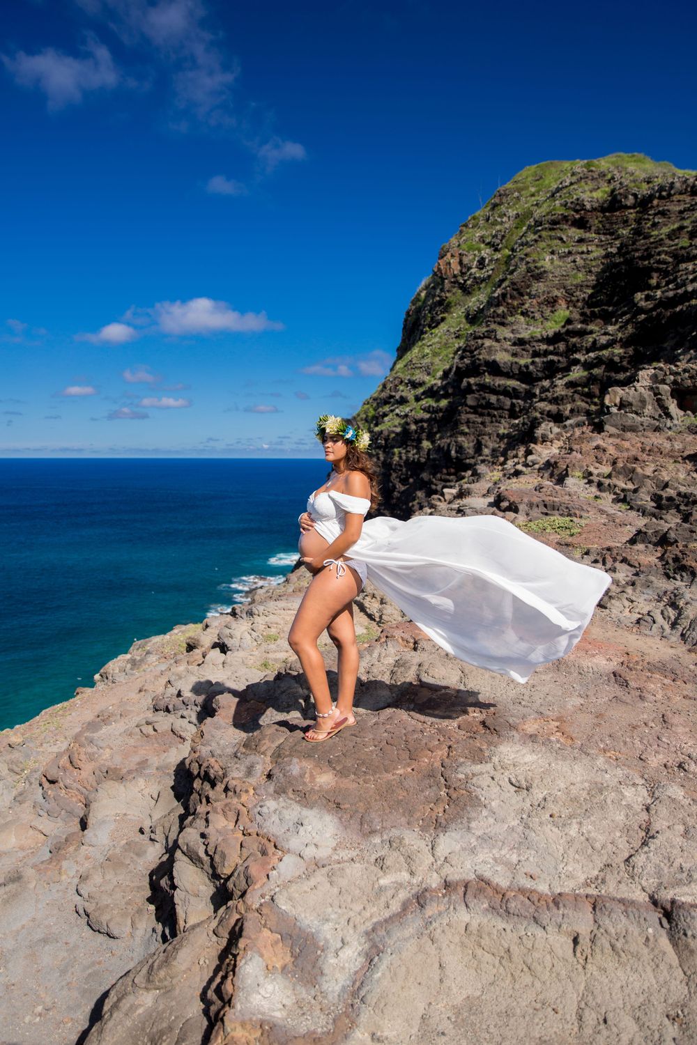 A maternity flowing white dress poses dramatically on a rocky cliff of Makapuʻu Lookout overlooking the bright blue Hawaiian ocean.