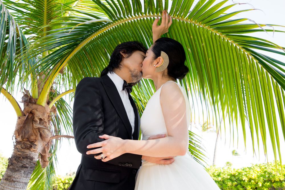 A romantic couple in wedding attire share a tender moment under a tropical palm frond at Waialae Beach in Kahala.