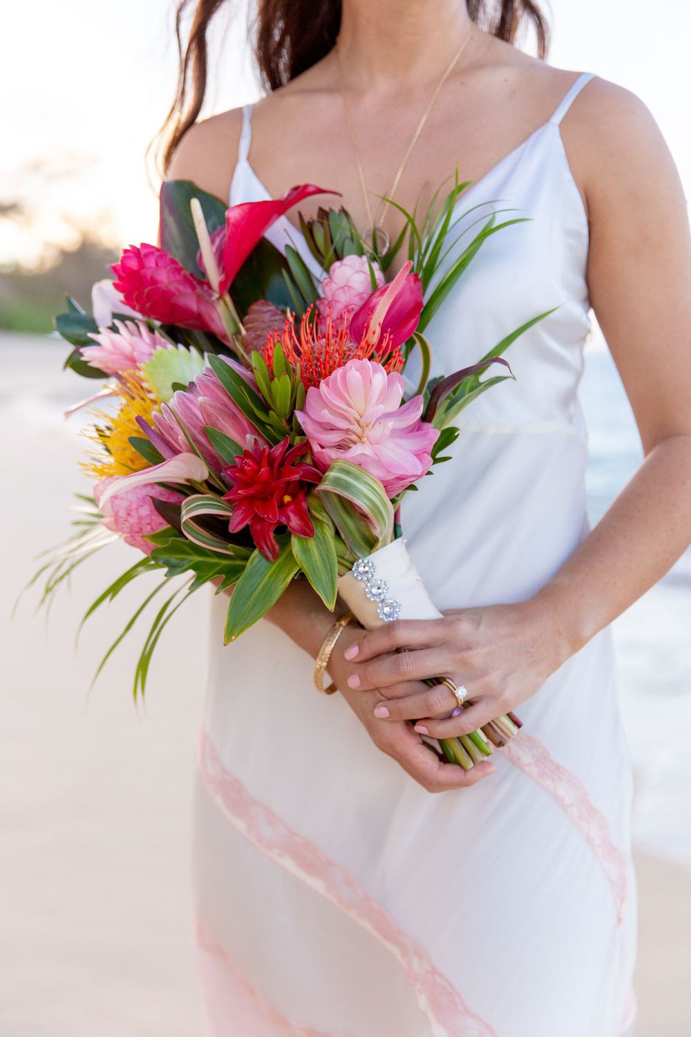 A vibrant tropical bouquet with pink and red flowers held against a white dress in soft sunlight.