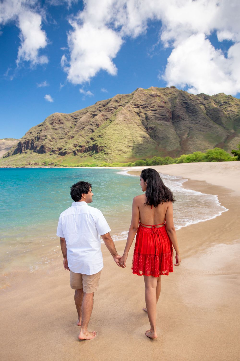 A couple walks hand in hand at scenic Makua Beach with turquoise water and mountains in the background.