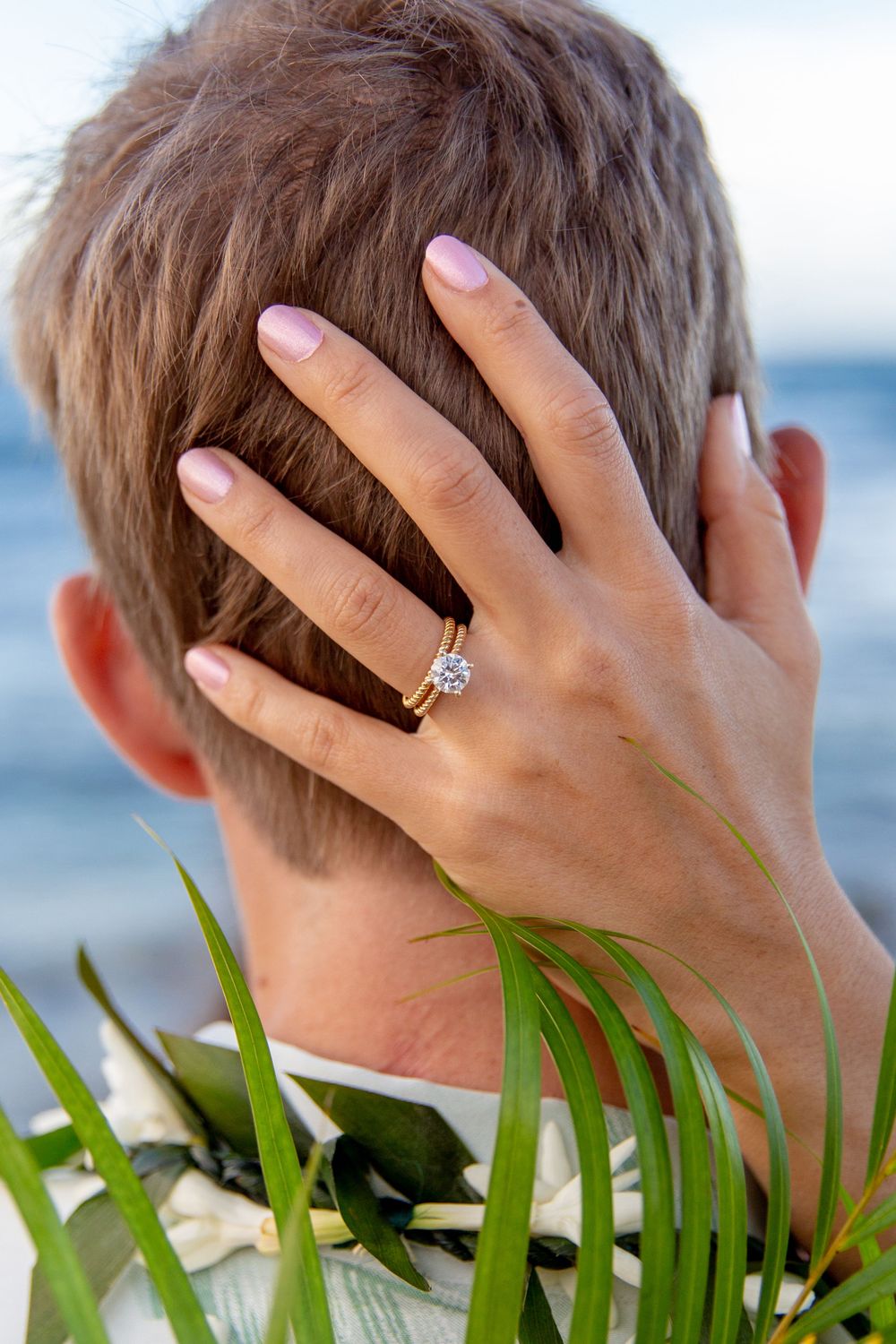A hand wearing an engagement ring rests on the back of a head near tropical foliage and ocean waters.