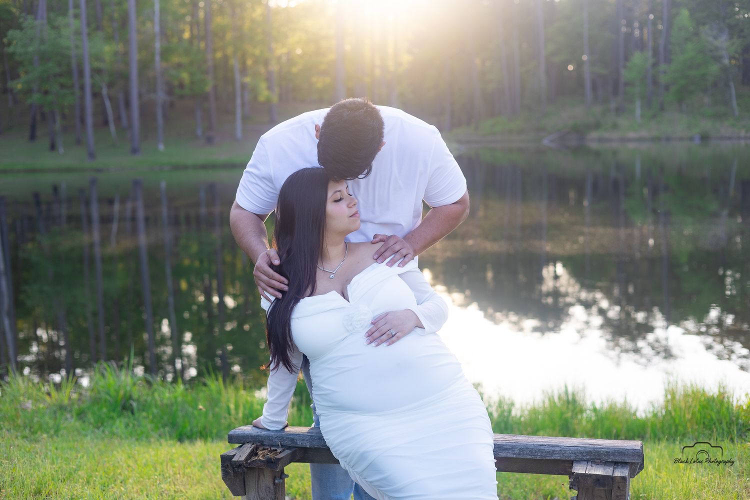 A couple shares a tender moment on a park bench near a tranquil lake at sunset.