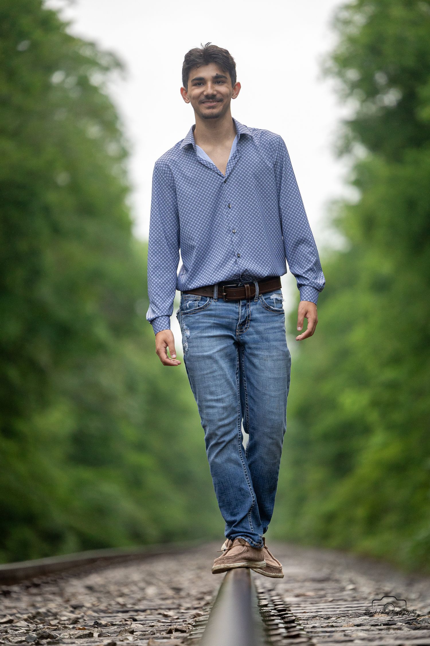 Person in blue polka dot shirt and jeans balancing while walking along train track rail in green forest setting.