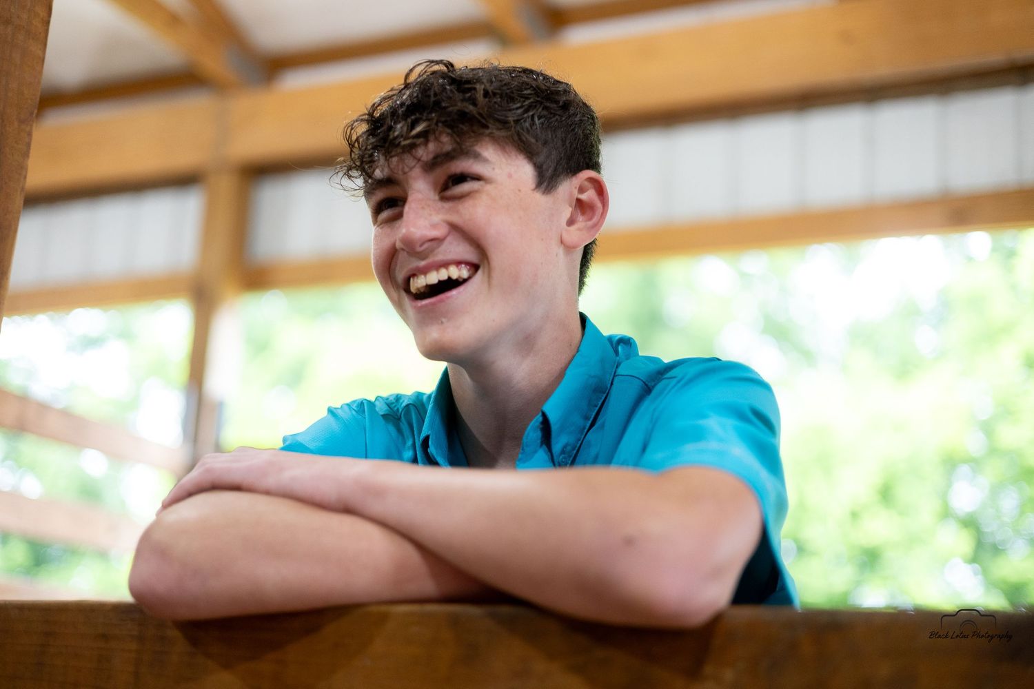 A young person in a bright blue shirt leans on a wooden railing while laughing joyfully in a sunlit space.