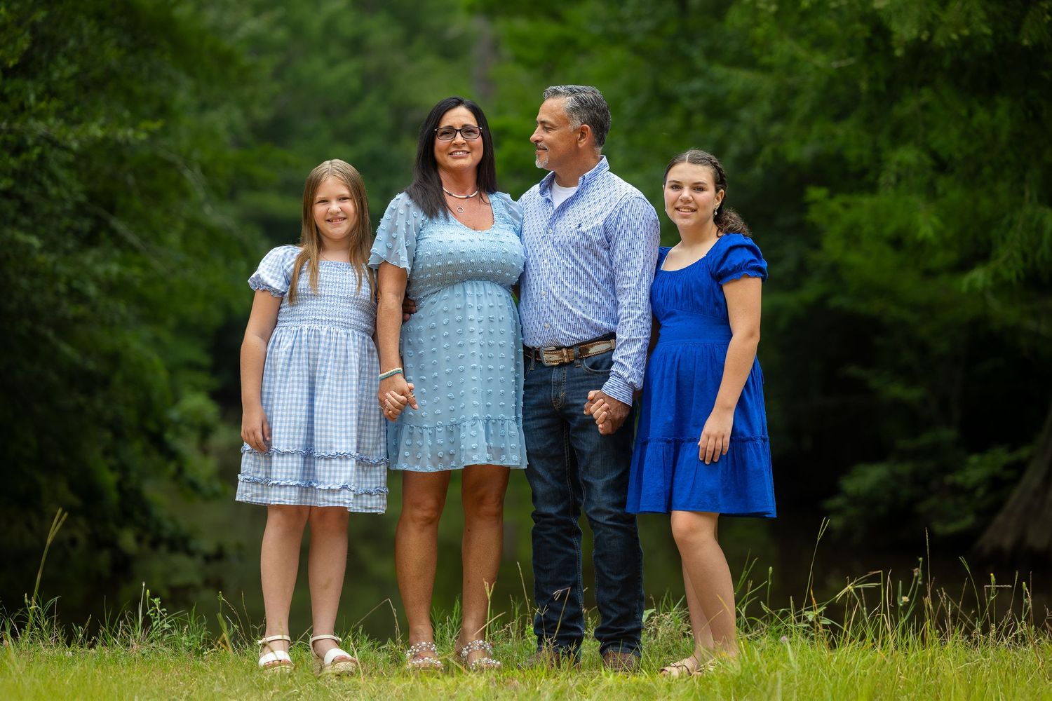 A family poses outdoors on a grassy hill with lush green trees in the background on a summer day.