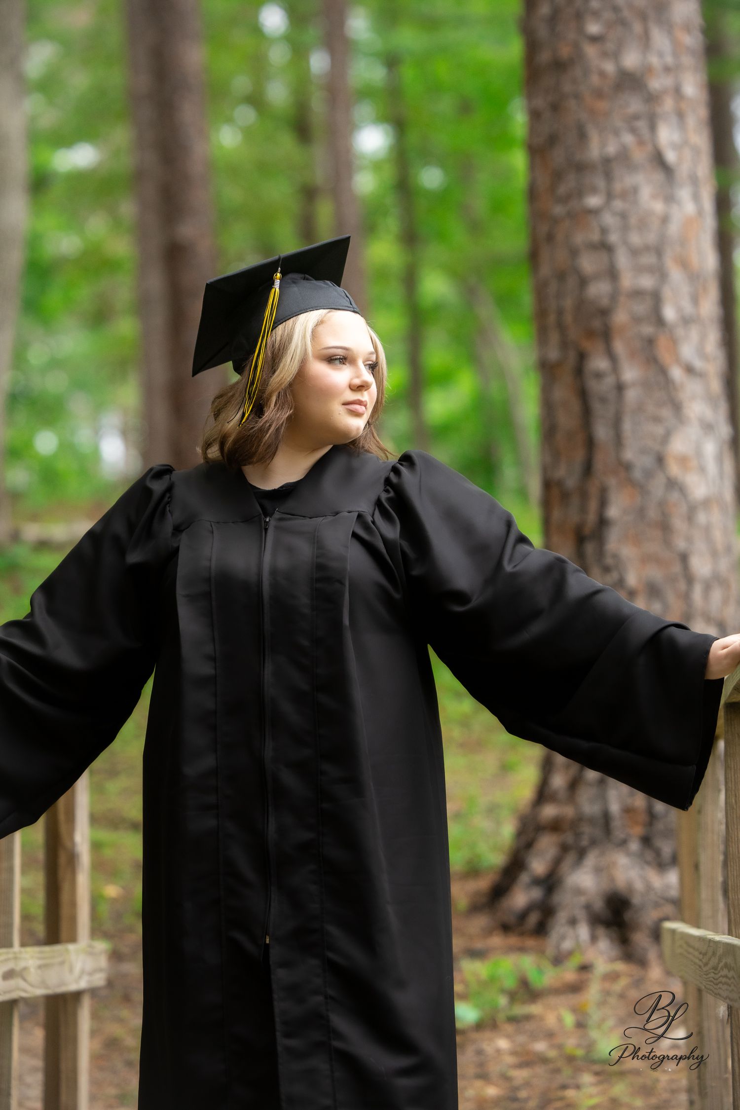 A young woman in black graduation cap and gown poses outdoors on a wooden bridge surrounded by tall pine trees.