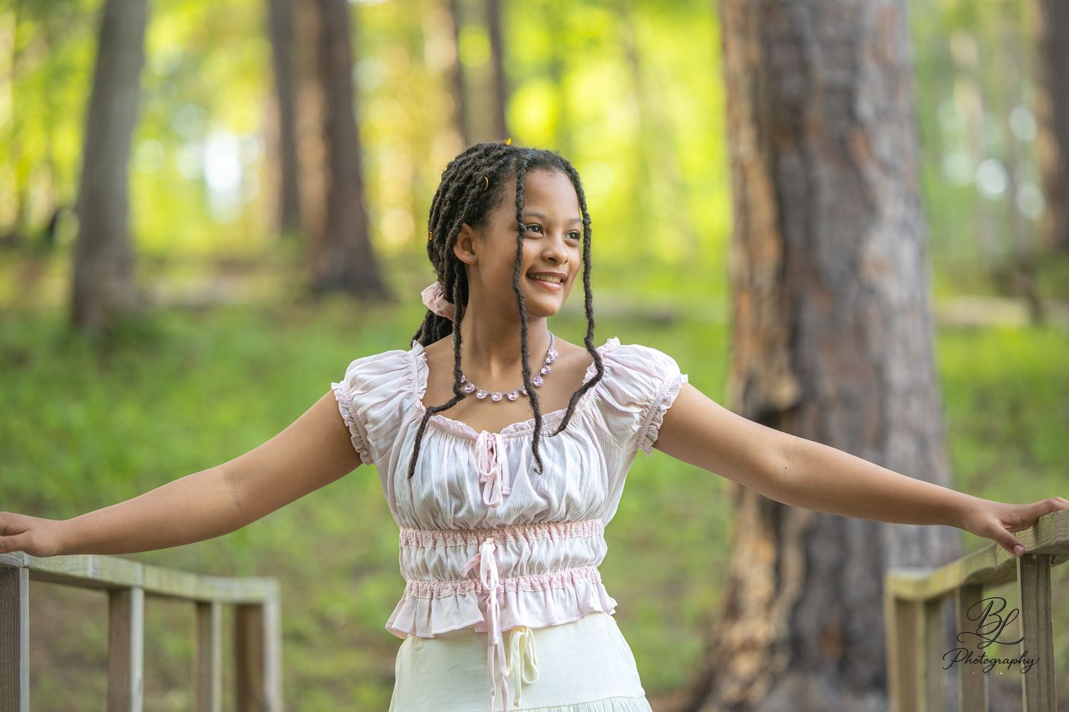 Young woman with braids smiling outdoors on a wooden bridge, arms outstretched, wearing a white top in a forest setting.
