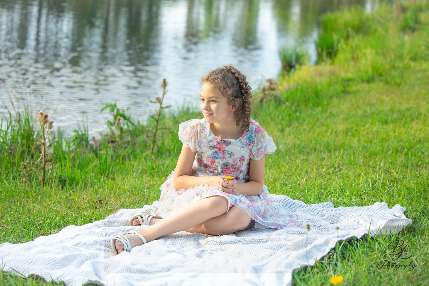 A young girl in a floral dress sits on a white blanket by a peaceful pond on a sunny day.