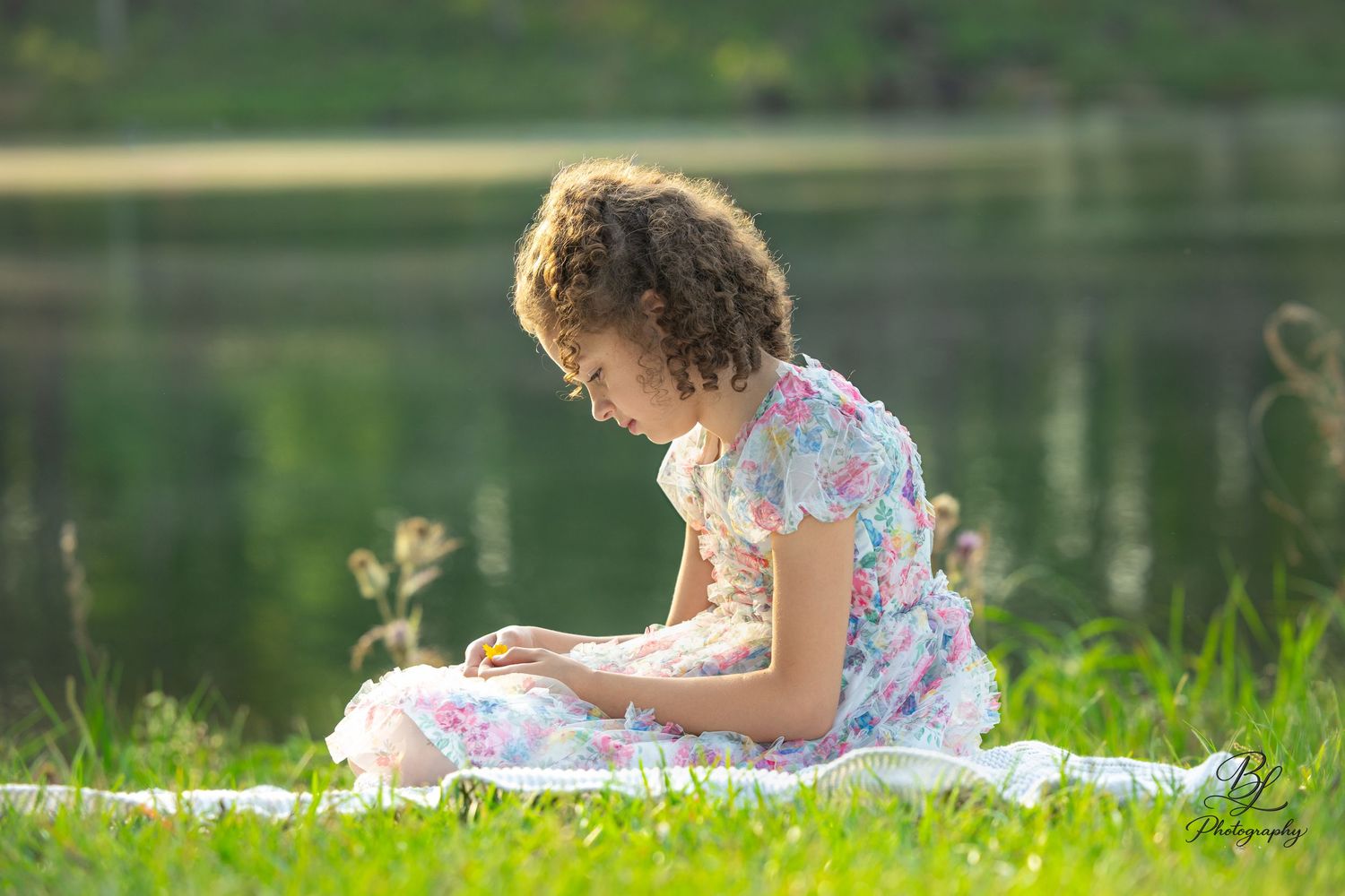 Curly-haired girl in floral dress sitting on blanket by lake reading in sunny green meadow.