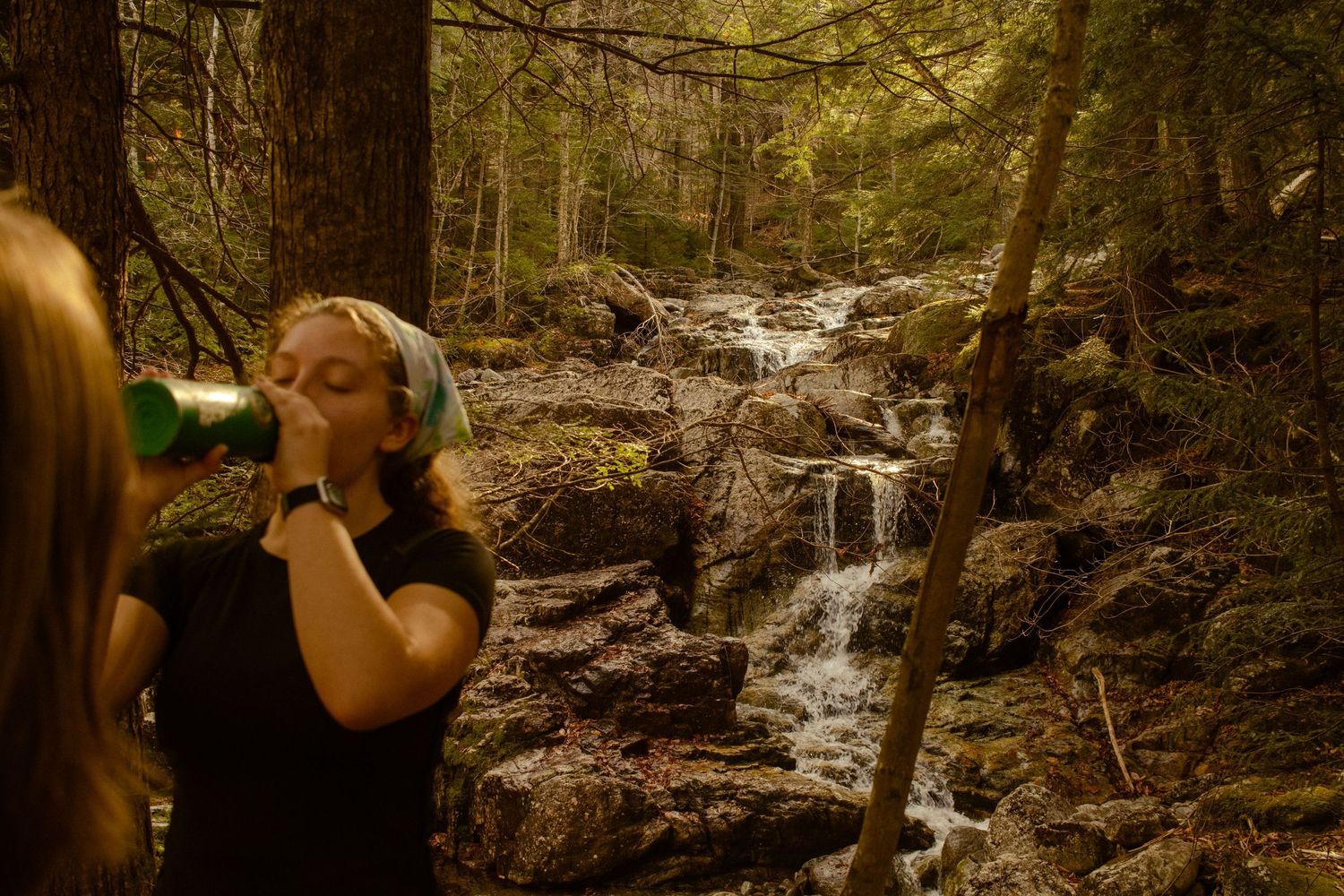 A girl drinking water next to a waterfall on the trail to the summit of Indian Head