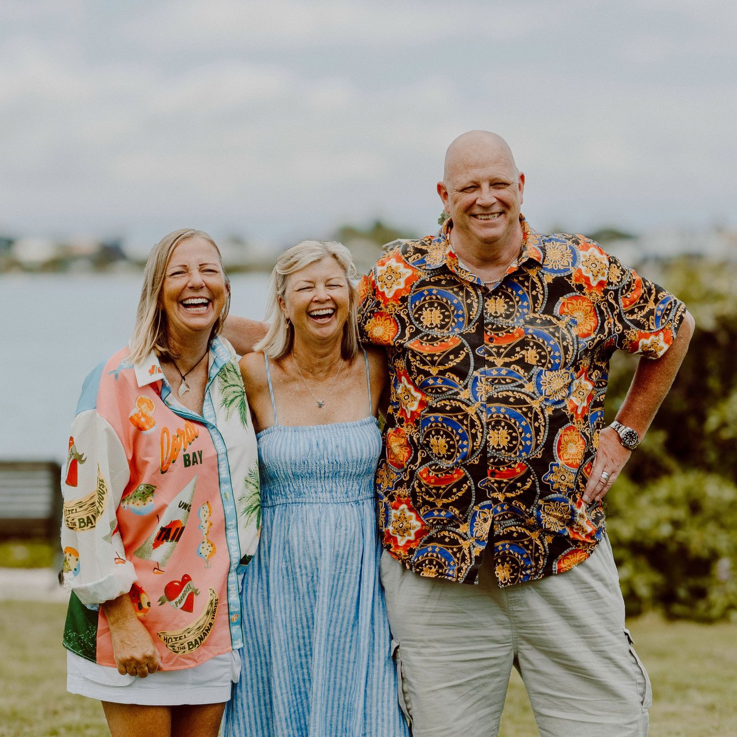 Three people wearing colorful shirts smile together by water on sunny day.