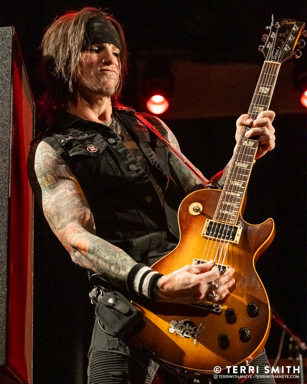 Ace Von Johnson of L.A. Guns plays guitar at the edge of the stage, leaning against a wall. Live concert in 2025.