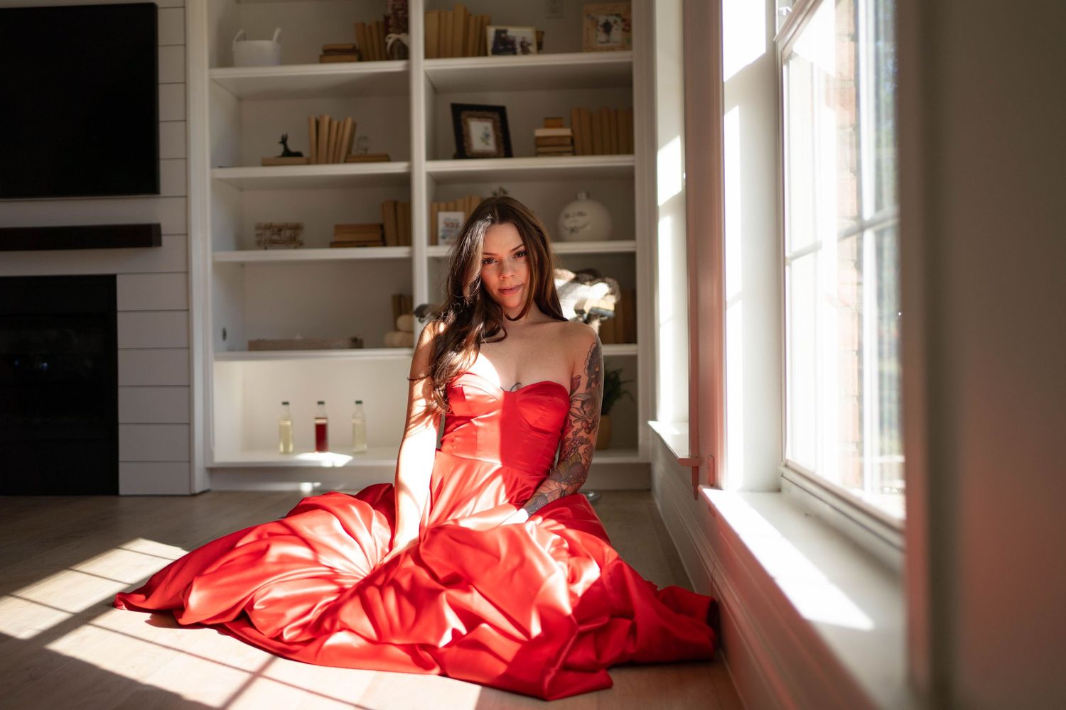 Woman in flowing red dress sits in sunlit window nook beside built-in shelving.