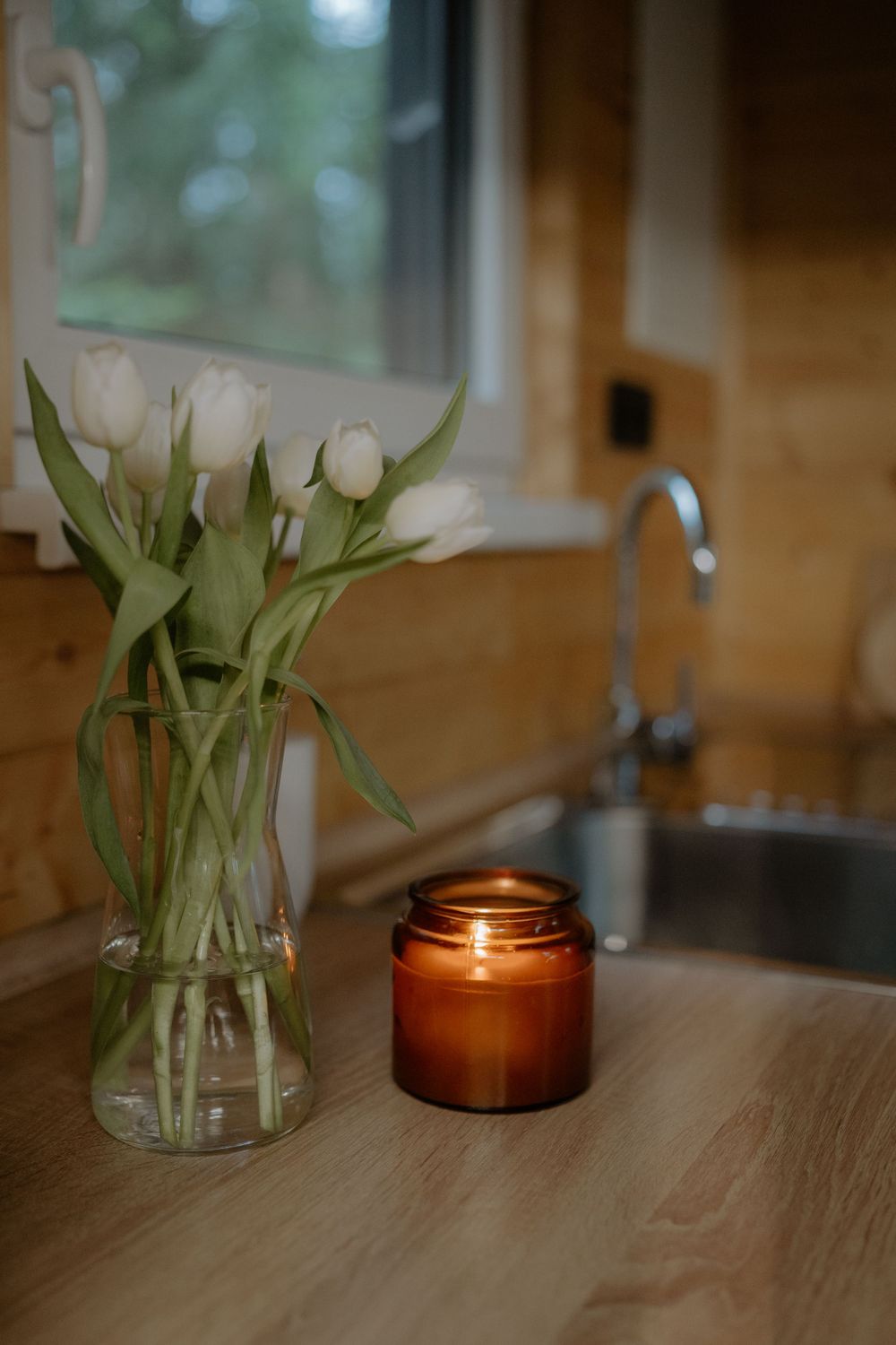 White tulips in clear vase next to amber candle on wooden countertop.
