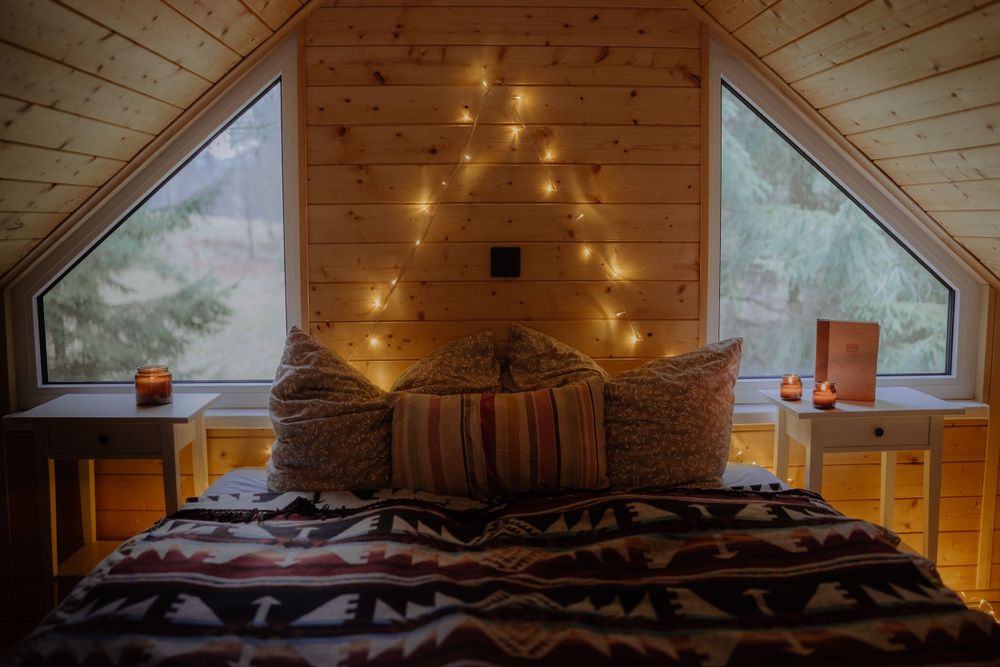 Cozy bed with string lights and triangle windows in wooden cabin loft.