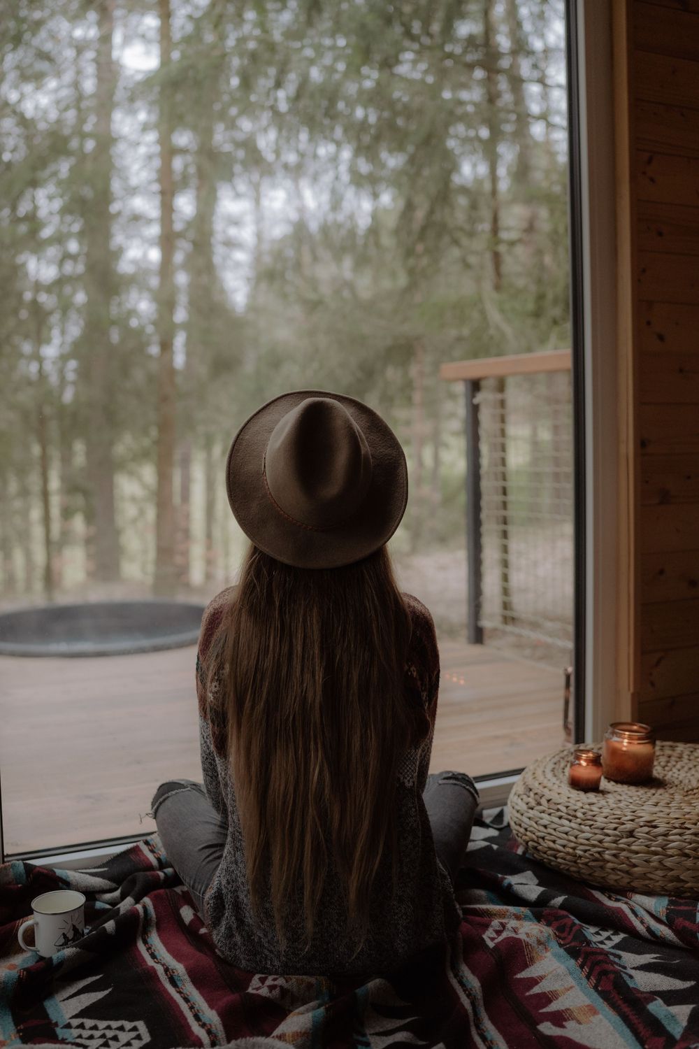 Person sitting by window in cozy cabin interior with blanket and forest view.