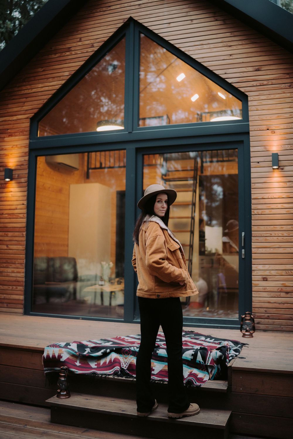 A-frame cabin exterior with large windows and person standing on wooden deck with blanket.