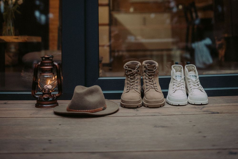 Vintage lantern, fedora hat, hiking boots and white sneakers arranged on a wooden floor by a window.