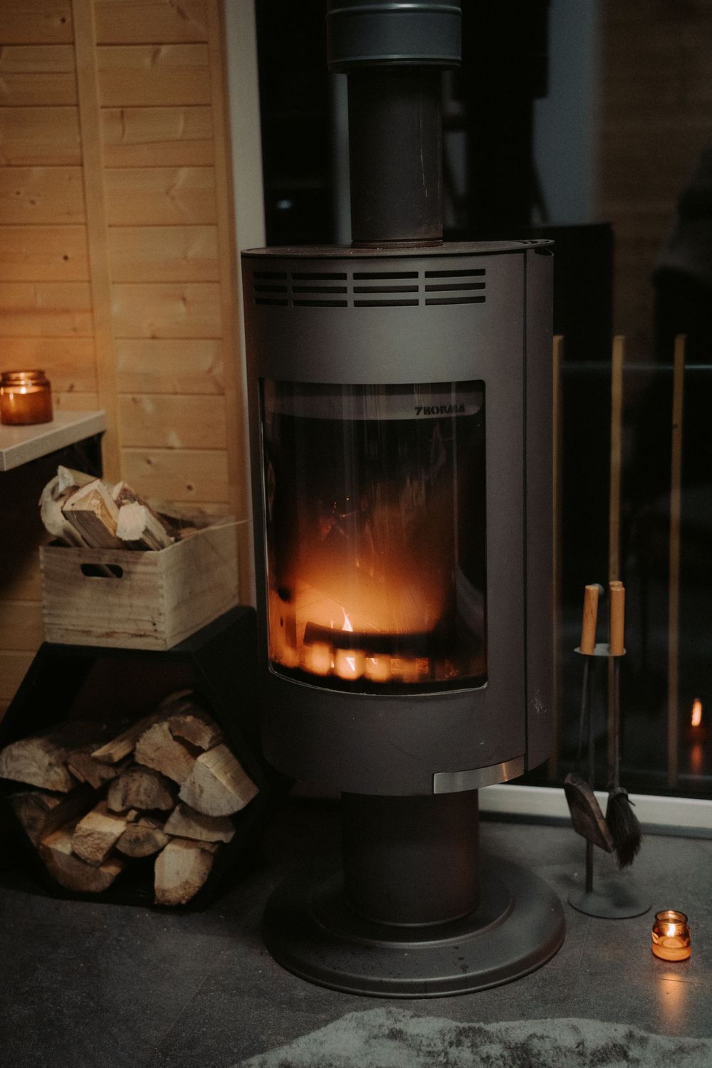 Modern wood burning stove with glowing fire and stacked firewood in corner.