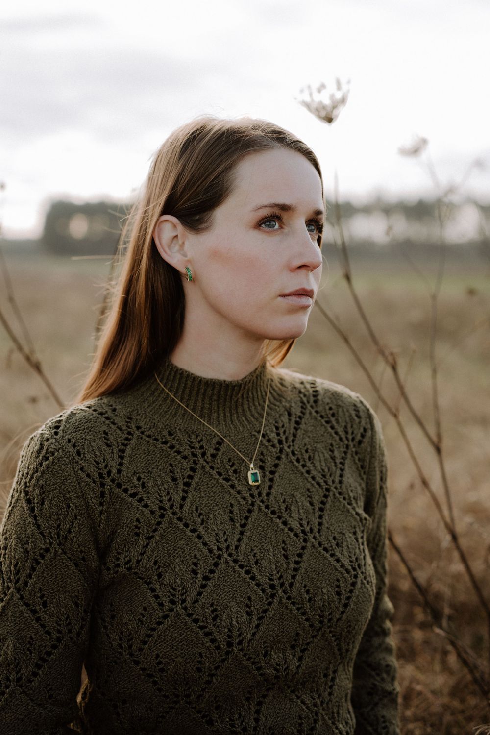 Person in olive knit sweater wearing emerald pendant necklace in autumn field.