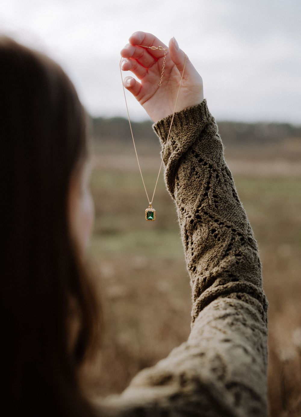 Hand holding delicate emerald pendant necklace against blurred outdoor background.