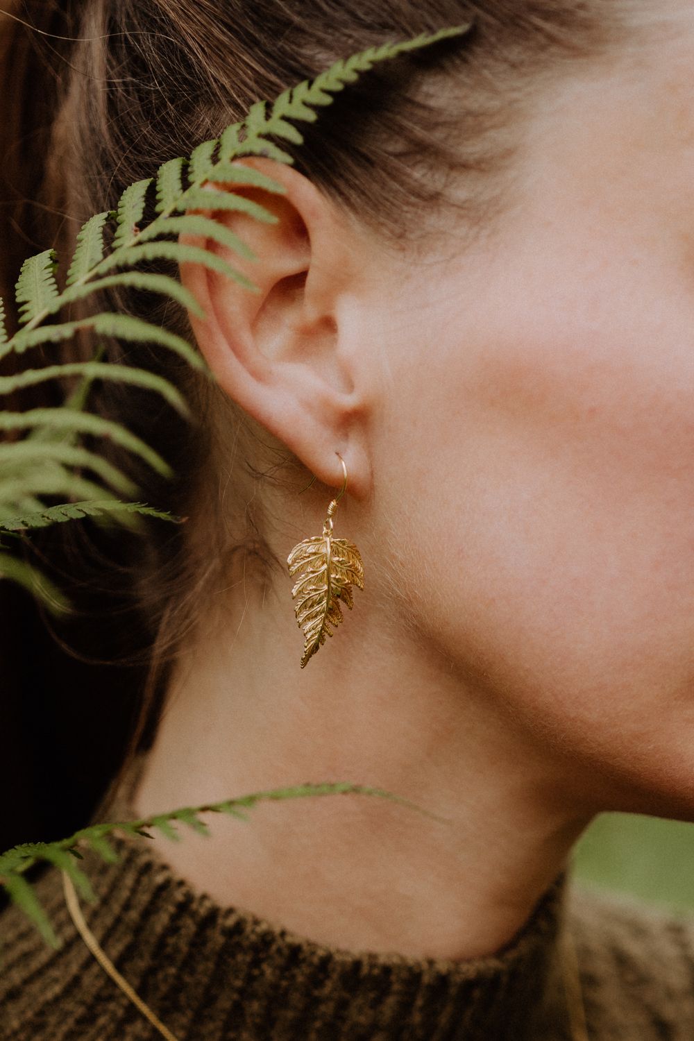 Gold fern leaf earring displayed against a backdrop of real green fern leaves.