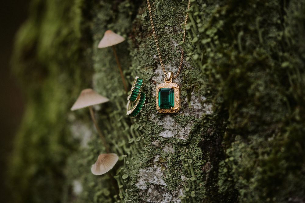 Emerald and gold jewelry earrings hang delicately against mossy tree bark with tiny mushrooms growing nearby.