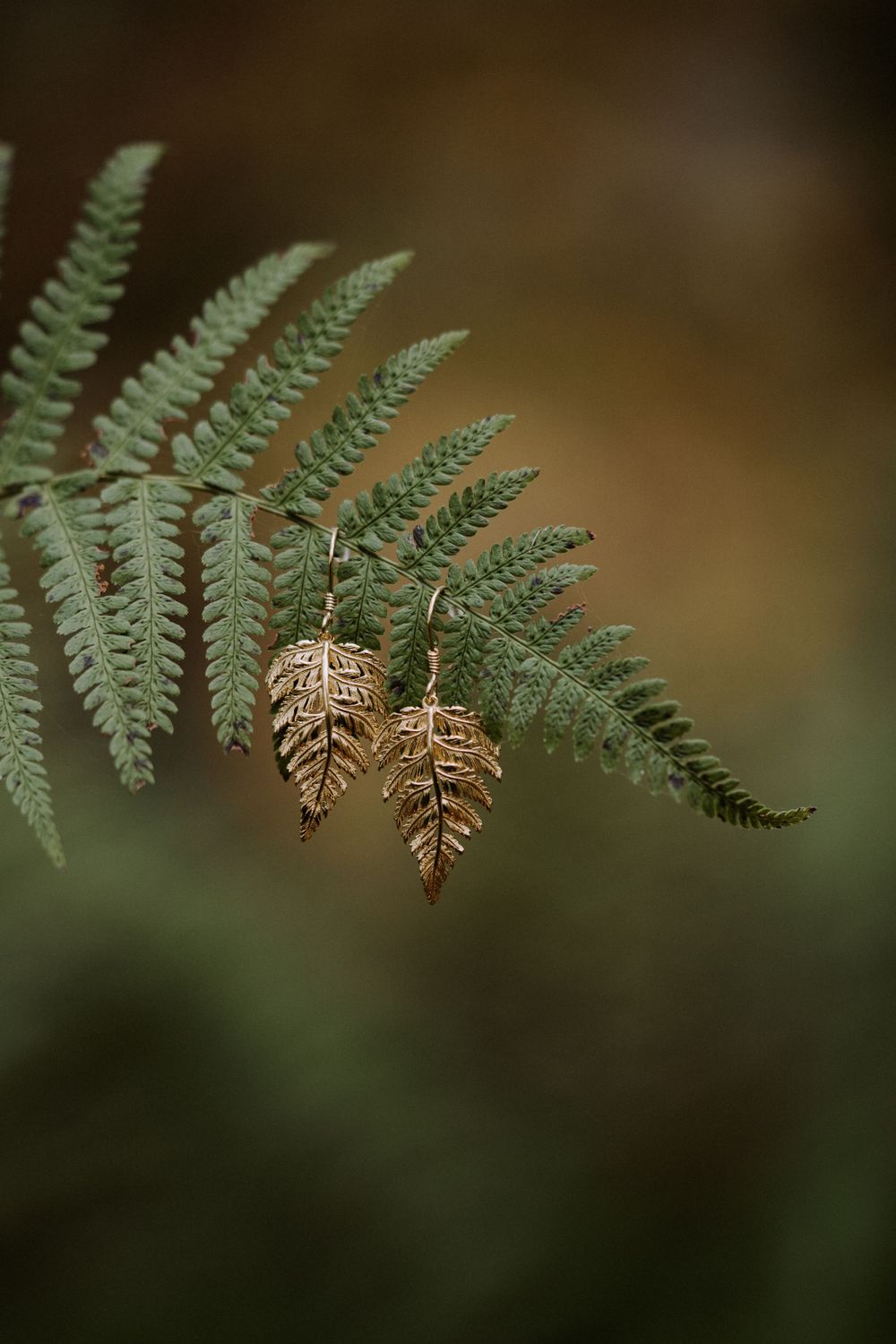 Pair of gold fern leaf earrings hanging against fresh green fern fronds.