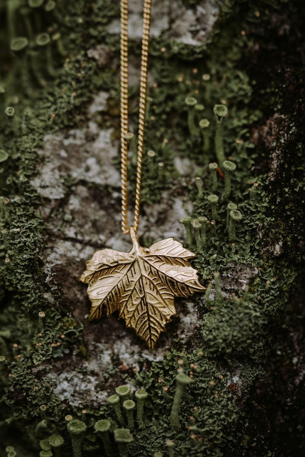 Gold maple leaf pendant necklace displayed on mossy tree bark surface.