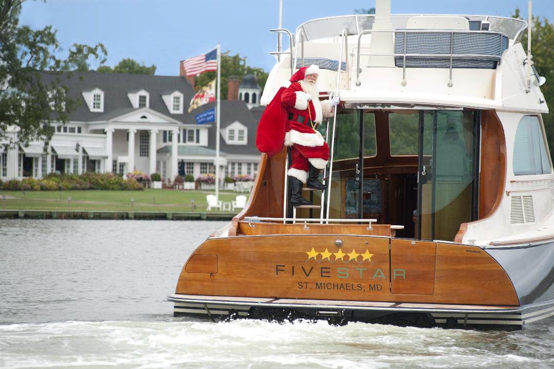 Santa Claus climbs aboard a luxury yacht with wooden trim on calm waters near waterfront homes.