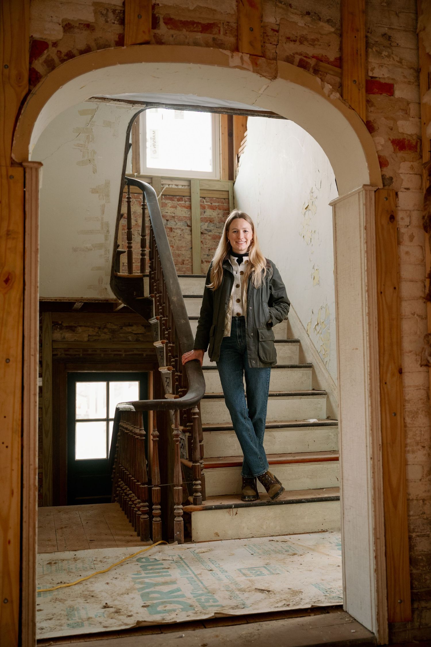 A figure stands on vintage wooden stairs framed by an arched doorway in a rustic brick interior.