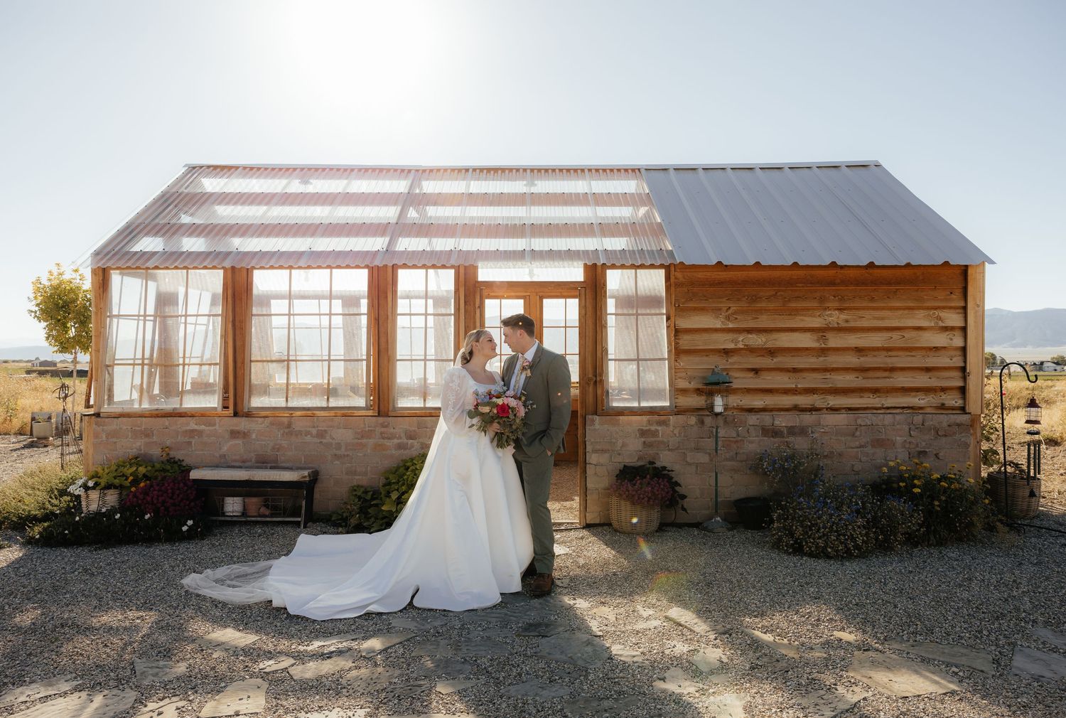 Bride and groom kissing in front of rustic wooden greenhouse on sunny wedding day.