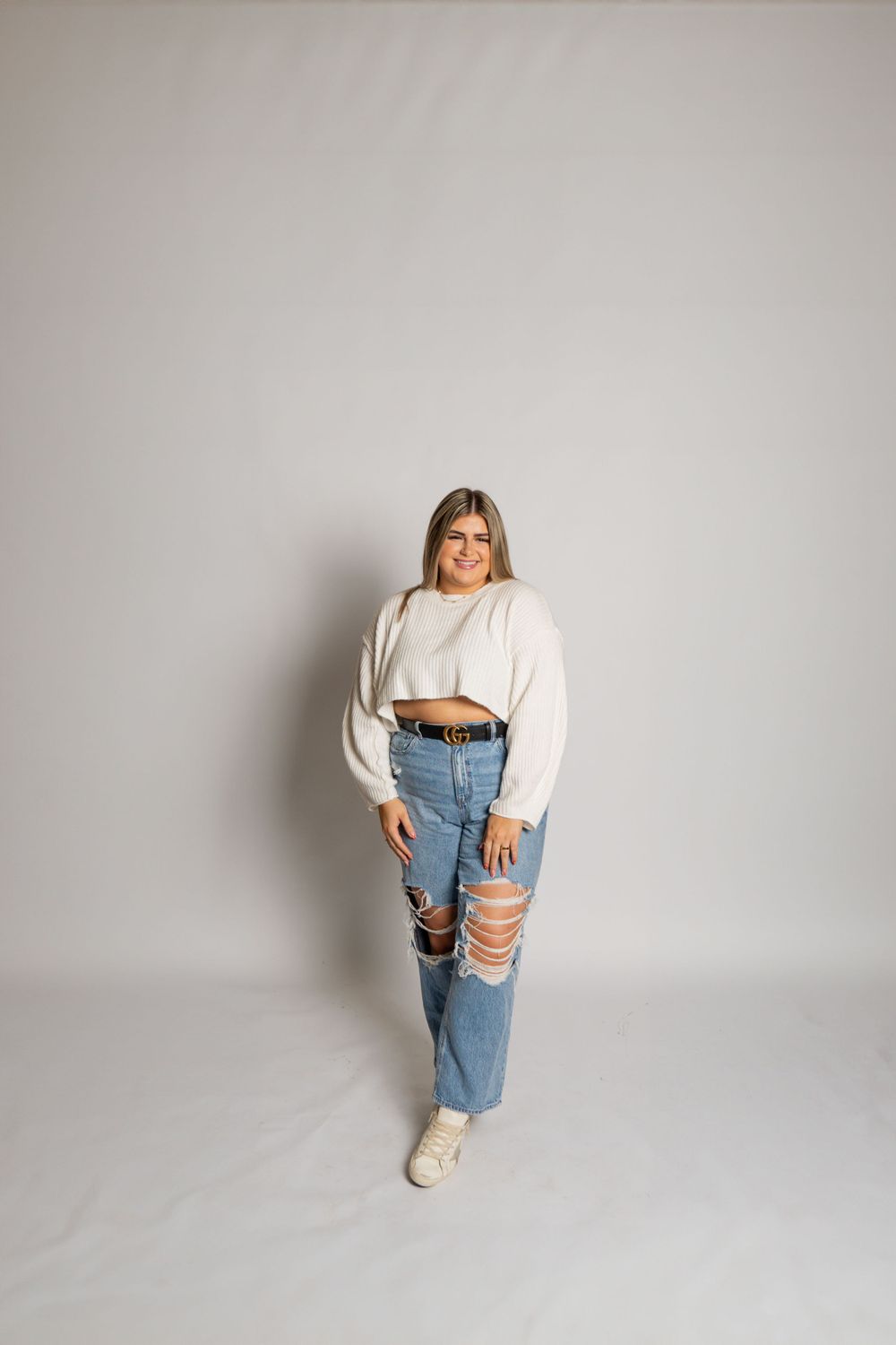 Woman in cropped sweater and distressed jeans poses against white studio background.