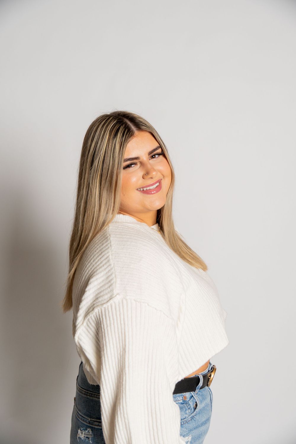 Woman in white knit sweater and jeans smiling against light gray background.