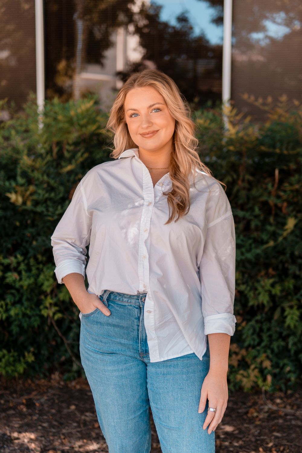 Casual portrait of someone wearing a classic white button-down shirt and blue jeans against a greenery background.