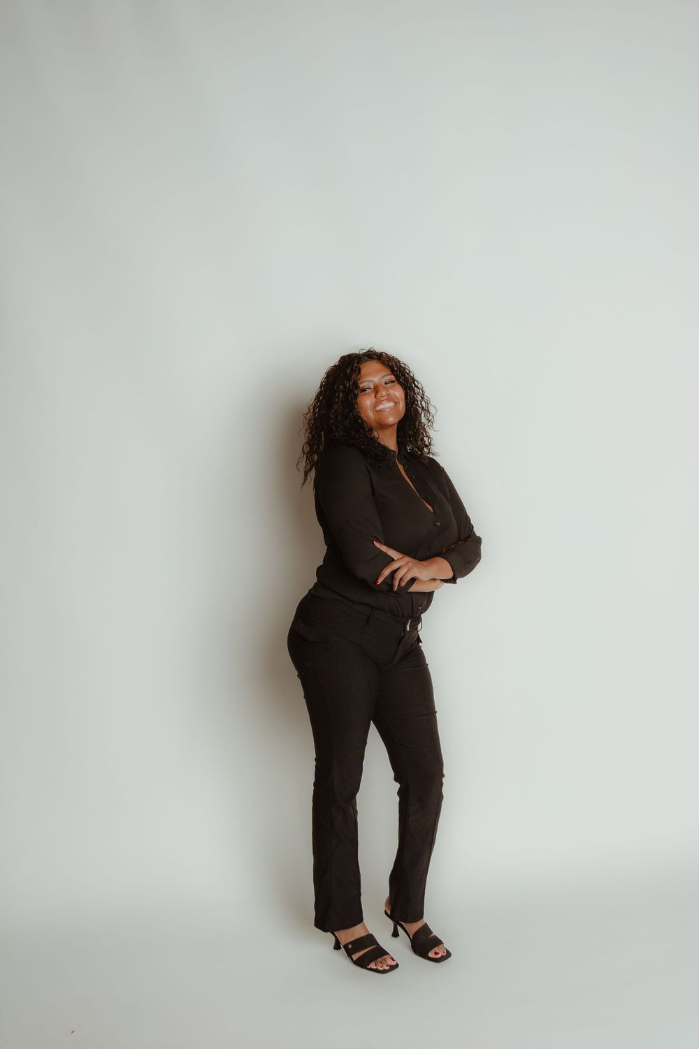 Woman in all black outfit poses confidently against white wall.
