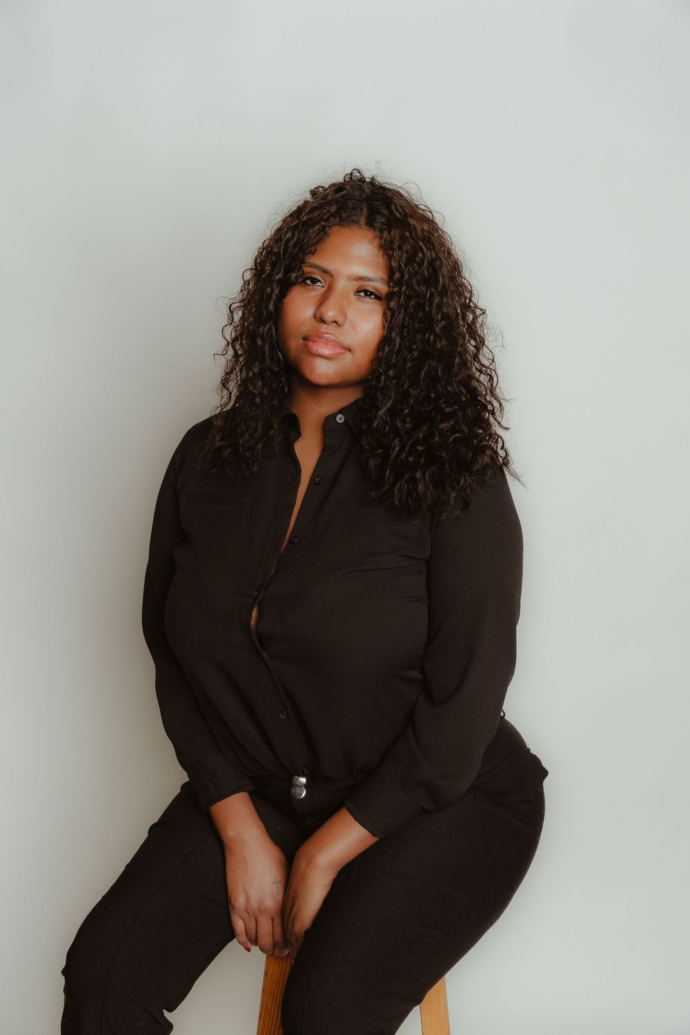 Woman with curly hair in black outfit sitting on stool against white background.