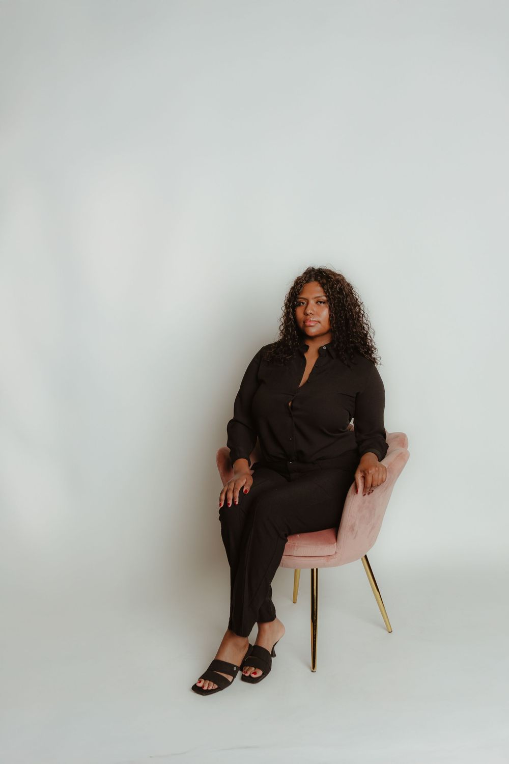 Woman in black ensemble seated elegantly on pink chair against white backdrop.