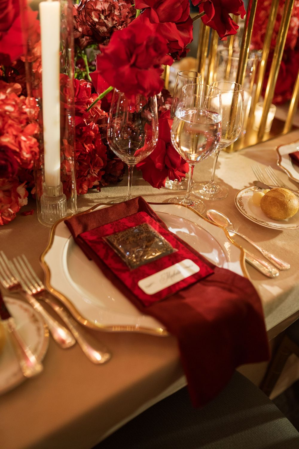 Elegant wedding table setting with red roses, gold cutlery, crystal glasses, and burgundy napkin on a white tablecloth.