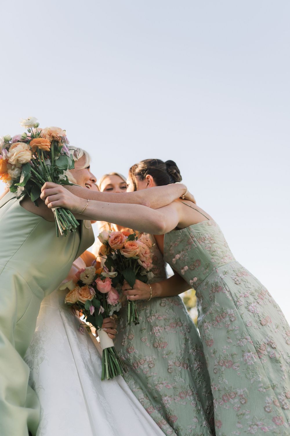 Bride and bridesmaids in green floral dresses share a group hug while holding peach bouquets against a clear sky.
