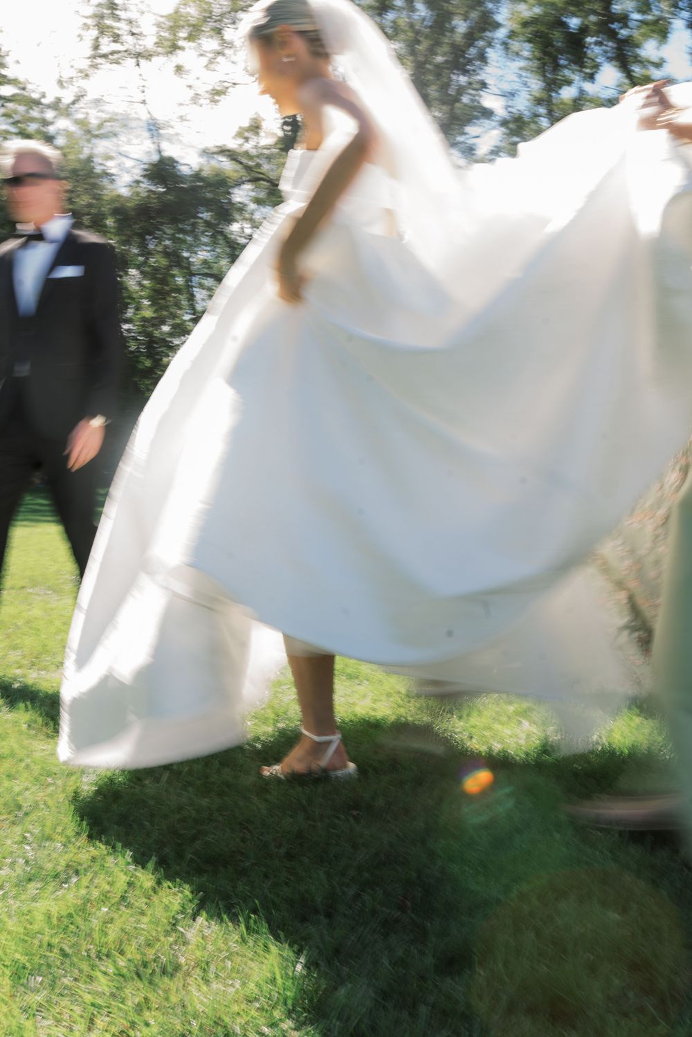 Bride in flowing white wedding gown and veil walks on grass outdoors with groom in tuxedo on a sunny day.