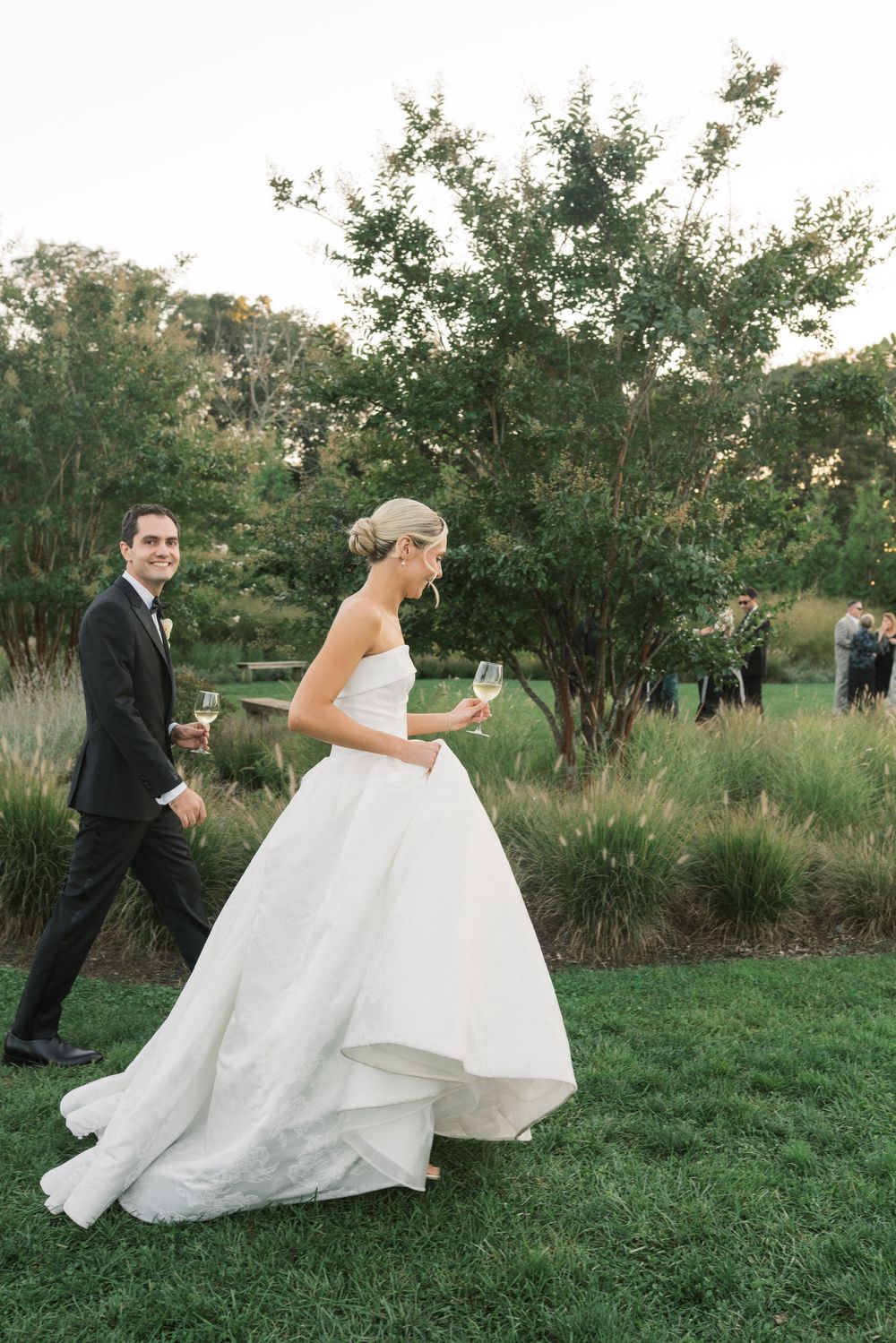 Bride and groom walk on green lawn holding champagne glasses at outdoor wedding reception.