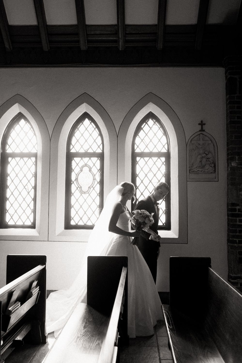 Black and white photo of bride and groom standing by gothic church windows, exchanging vows amid wooden pews.