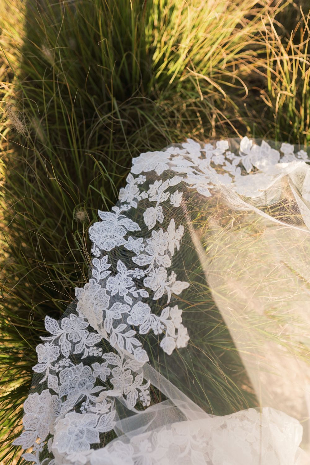Close-up of delicate white floral lace wedding veil draped over green grass in soft natural sunlight.