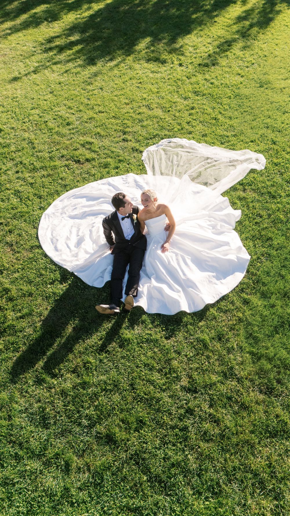 Aerial drone photo of bride and groom lying on green grass, wedding dress spread wide, looking at each other on their wedding day.