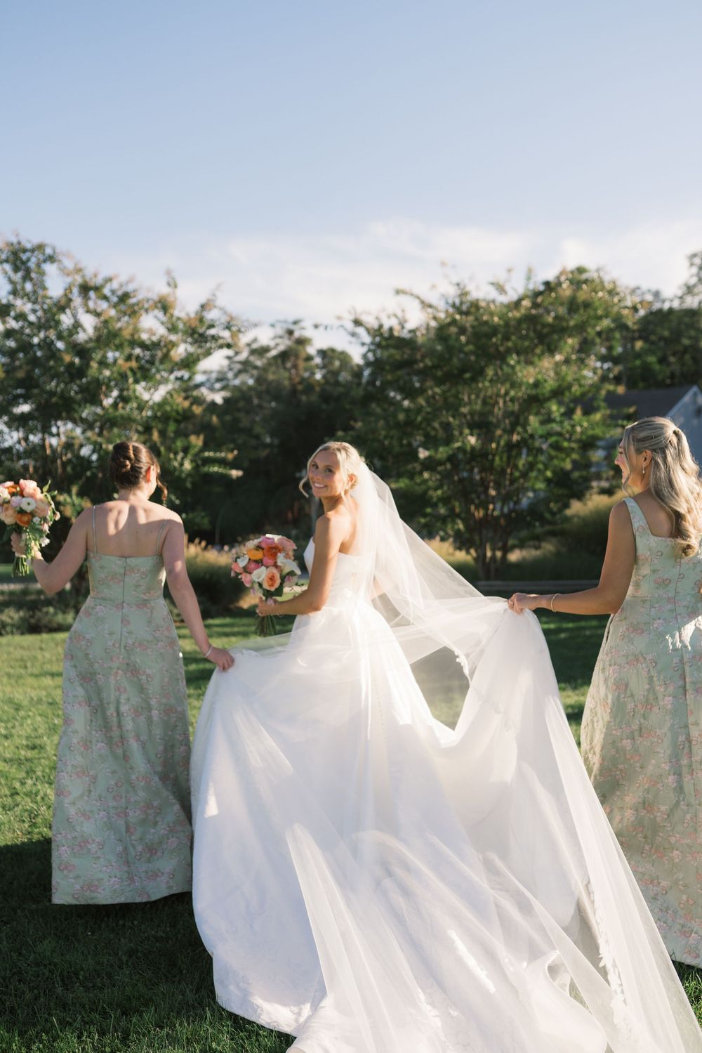 Bride in white gown and veil walks with bridesmaids in floral dresses on green lawn.