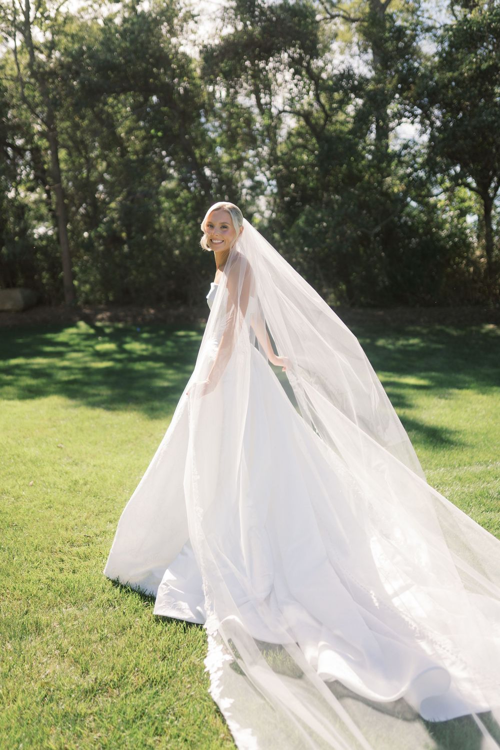 Bride in flowing white gown and cathedral veil smiles outdoors on lush green lawn surrounded by trees.