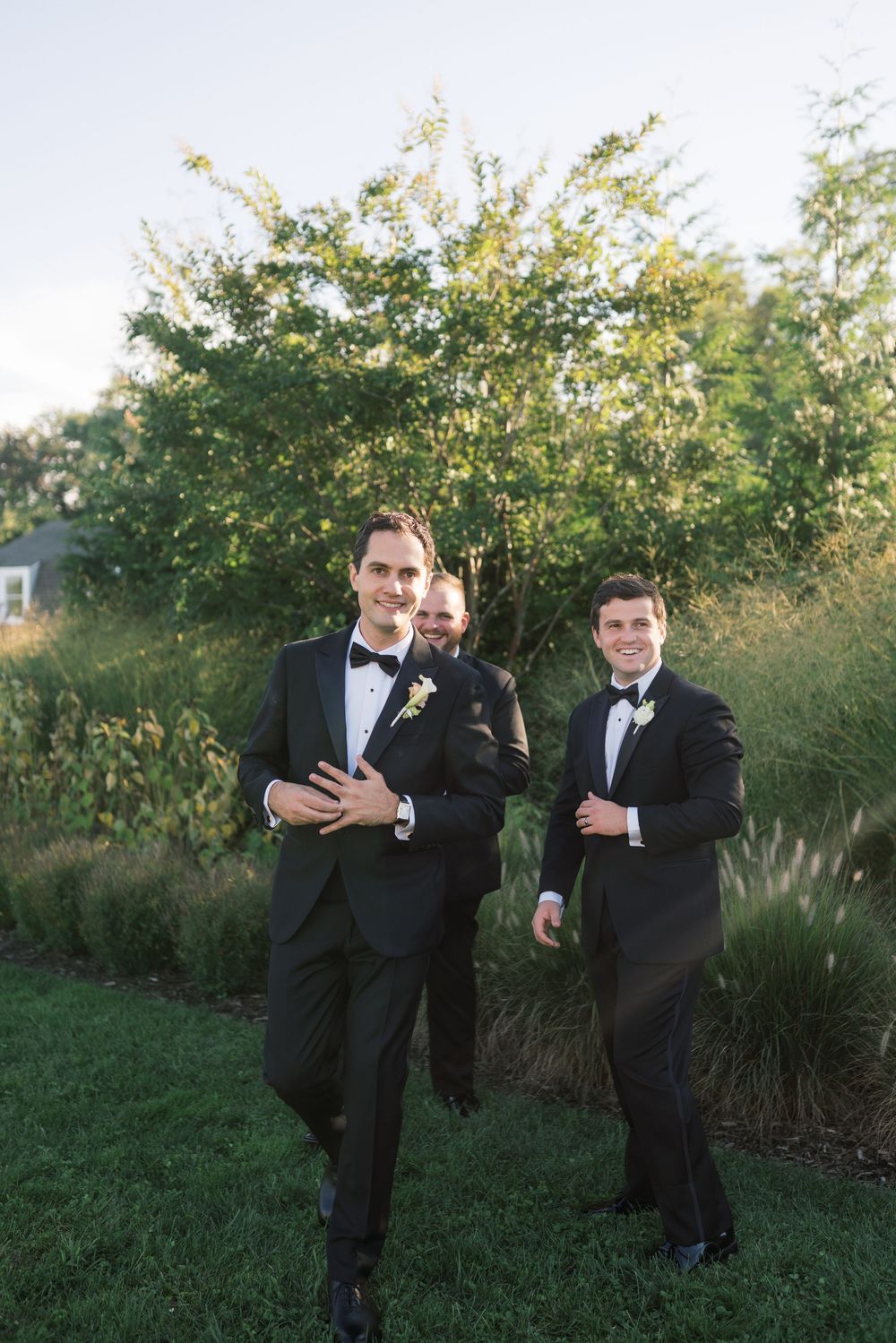 Three groomsmen in black tuxedos with boutonnieres pose outdoors on a lush green lawn at a wedding.