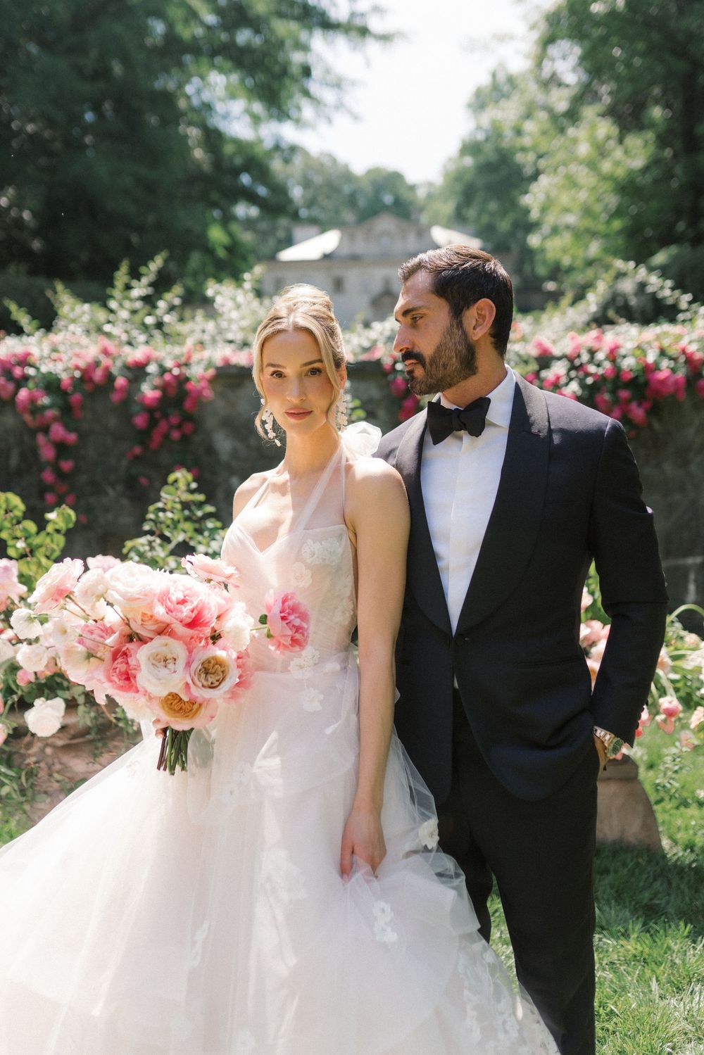 Bride in white gown holding pink bouquet stands with groom in black tuxedo in a garden with roses.