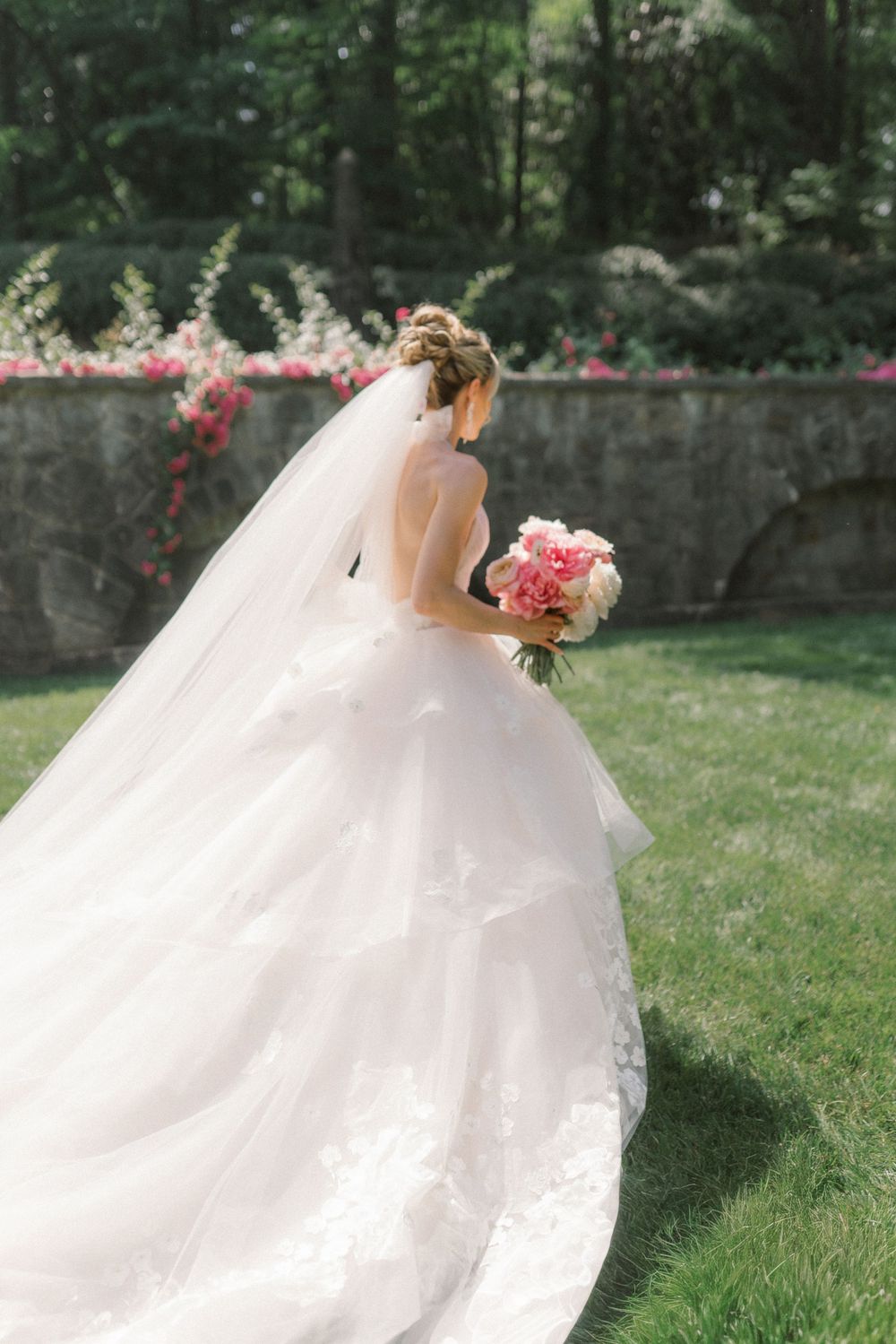 Bride in a flowing white ballgown with veil holds pink bouquet outdoors near stone wall with blooming flowers.